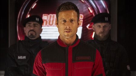 Glen Powell with a red outfit on staring directly ahead in The Running Man