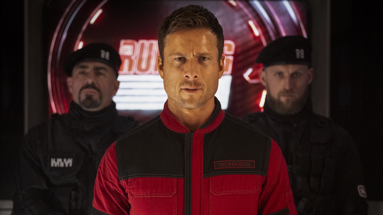 Glen Powell with a red outfit on staring directly ahead in The Running Man