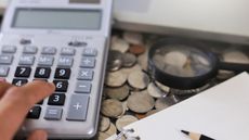 A person's fingers work on a calculator that sits atop a pile of coins next to a magnifying glass.