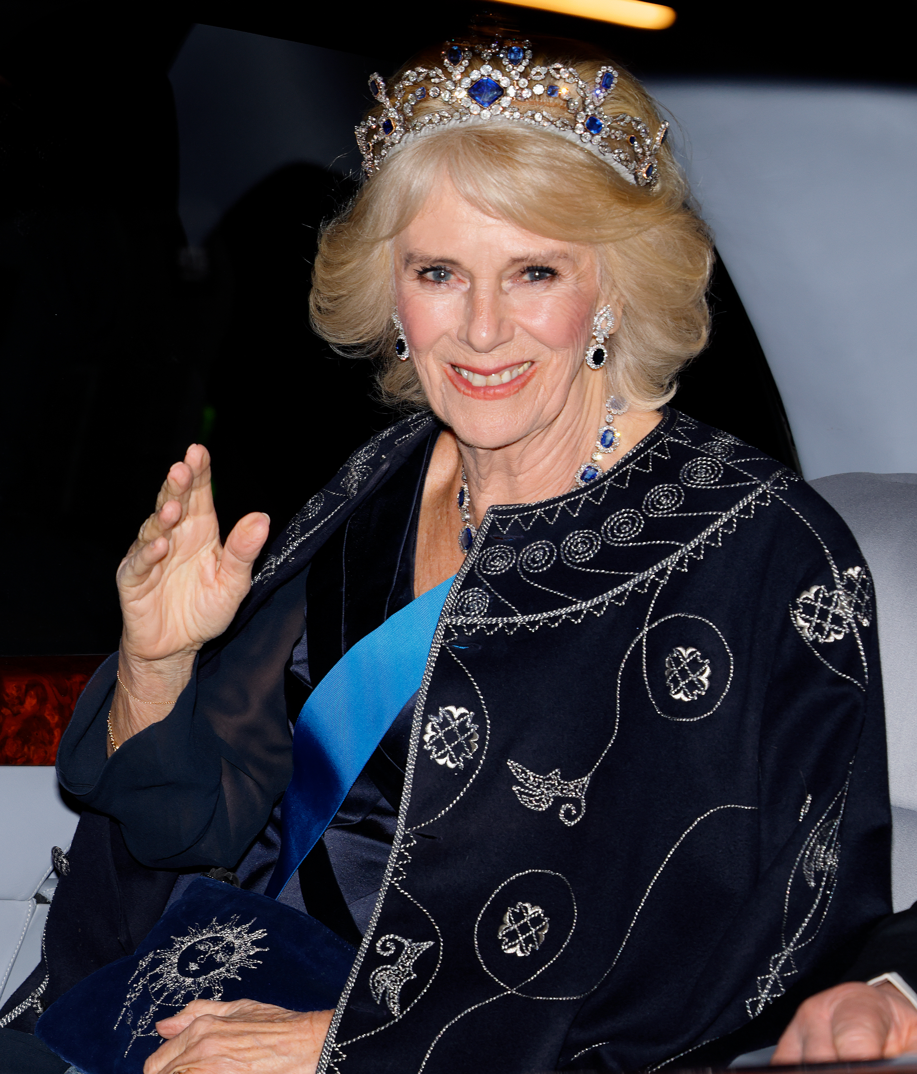 Queen Camilla wearing a blue robe and diamond and sapphire tiara waving from a car