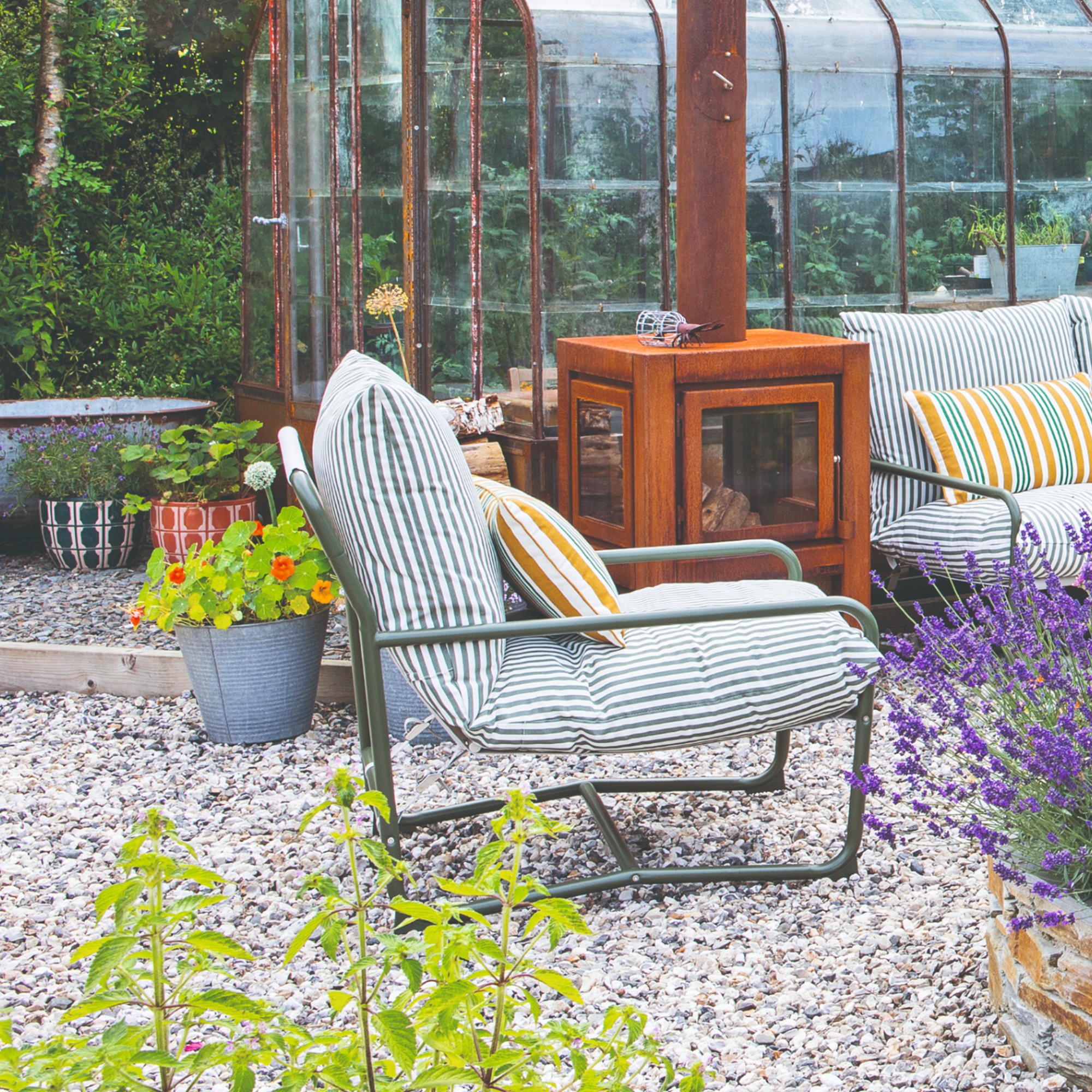 A gravel garden with a greenhouse and the John Lewis Marcy garden chair in green stripe with a matching two-seater outdoor sofa