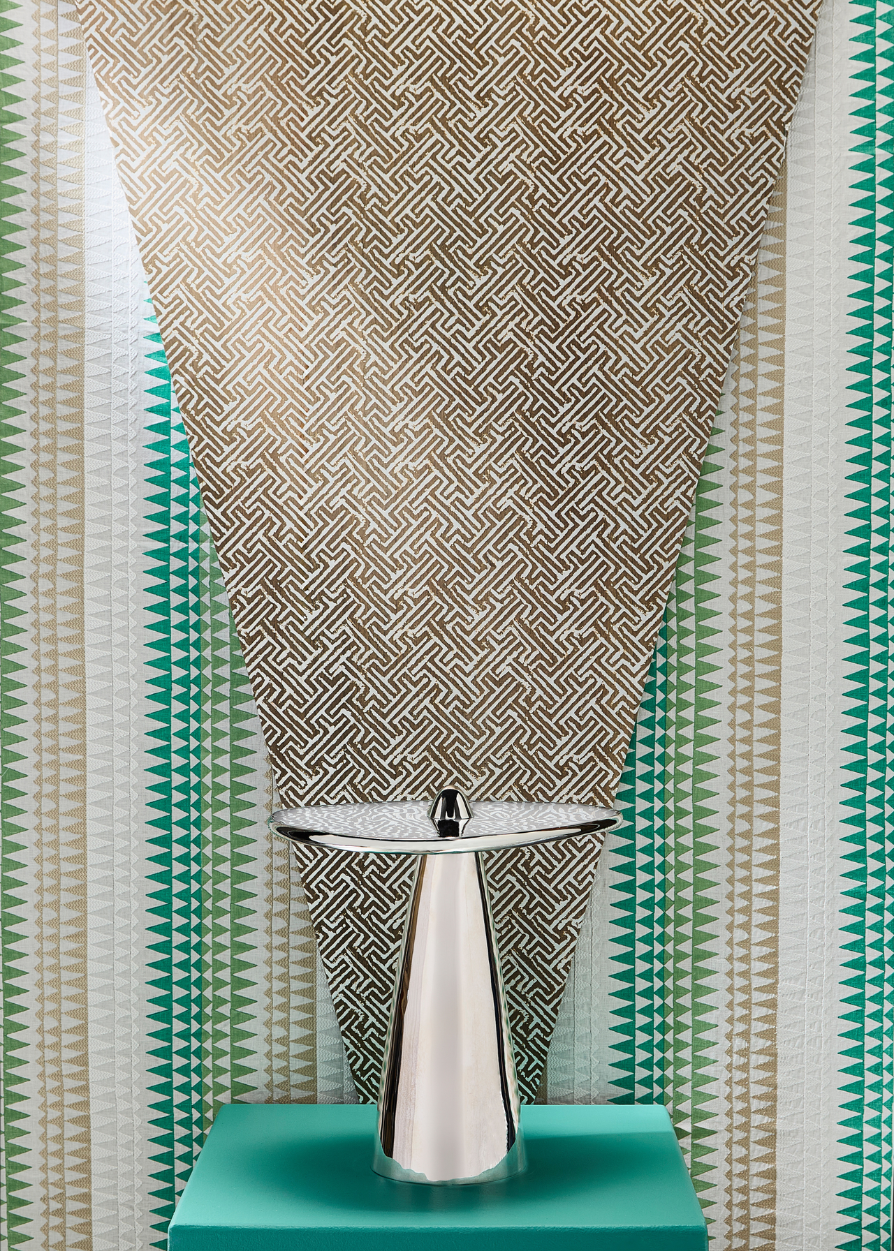 a design showcase featuring a metal side table sitting on top of a green pedestal, where the base of the table is pointing up and through the centre of the round top, in front of a green beige and white patterned wallpaper behind with a gold patterned material laid over the top in a triangle shape pointing down towards the table from behind it