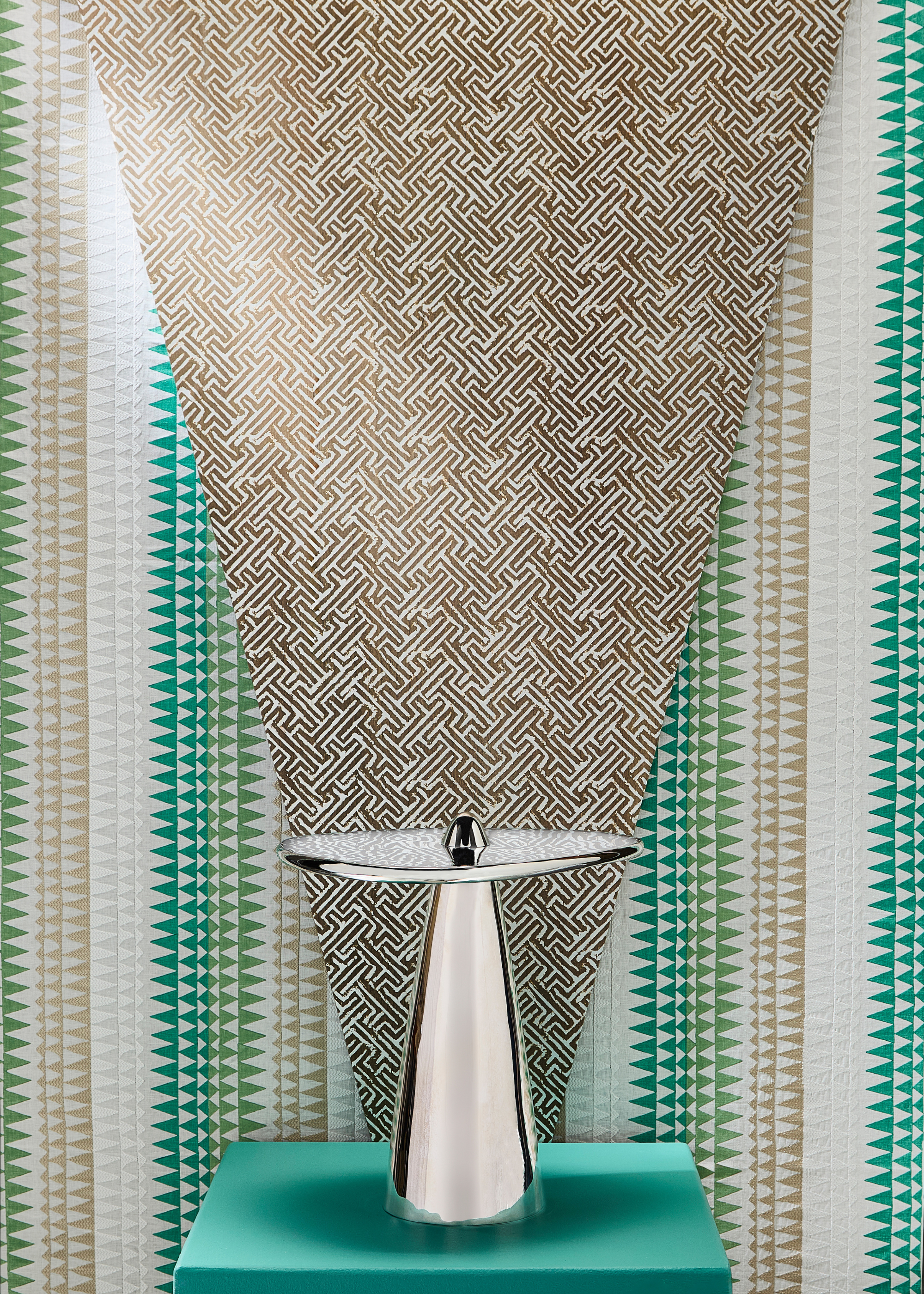 a design showcase featuring a metal side table sitting on top of a green pedestal, where the base of the table is pointing up and through the centre of the round top, in front of a green beige and white patterned wallpaper behind with a gold patterned material laid over the top in a triangle shape pointing down towards the table from behind it