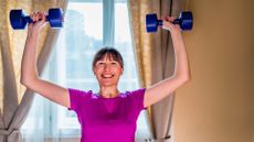 woman wearing a pink tshirt pressing two blue dumbbells overhead and smiling. there's a curtained window and plain wall behind her.