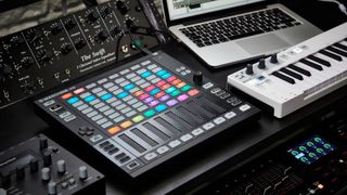 Lots of music gear on a desk including synthesizer, groovebox, MIDI controller, and more