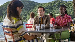 Lexi (Ronni Ancona) sits at a cafe table on the beach with her back to the camera. On the other side of the table sit Neville (Ralf Little) and Naomi (Shantol Jackson), who are scrutinising her