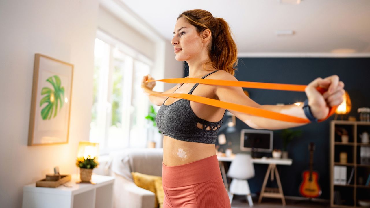 woman standing in a living room holding an orange resistance band in both hands and pulling it apart at chest height. 