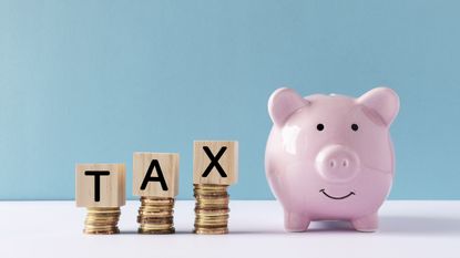 The word tax is spelled out on blocks balanced on top of successively higher stacks of coins next to a piggy bank.