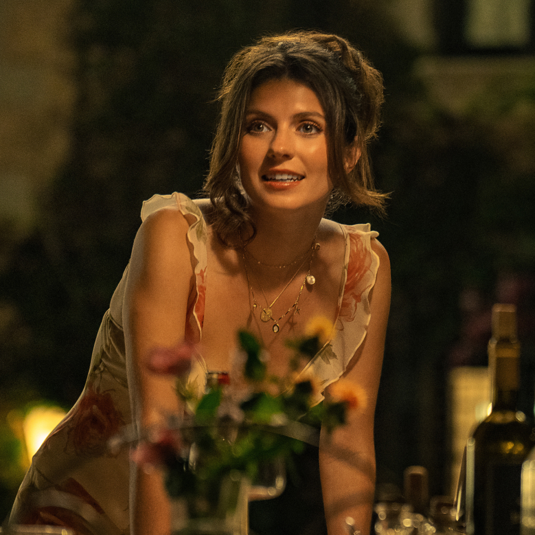 A woman (Emily Bader as Poppy) stands while resting both hands on an outdoor table decorated with wildflowers and bottles, in the Netflix movie 'People We Meet on Vacation.'