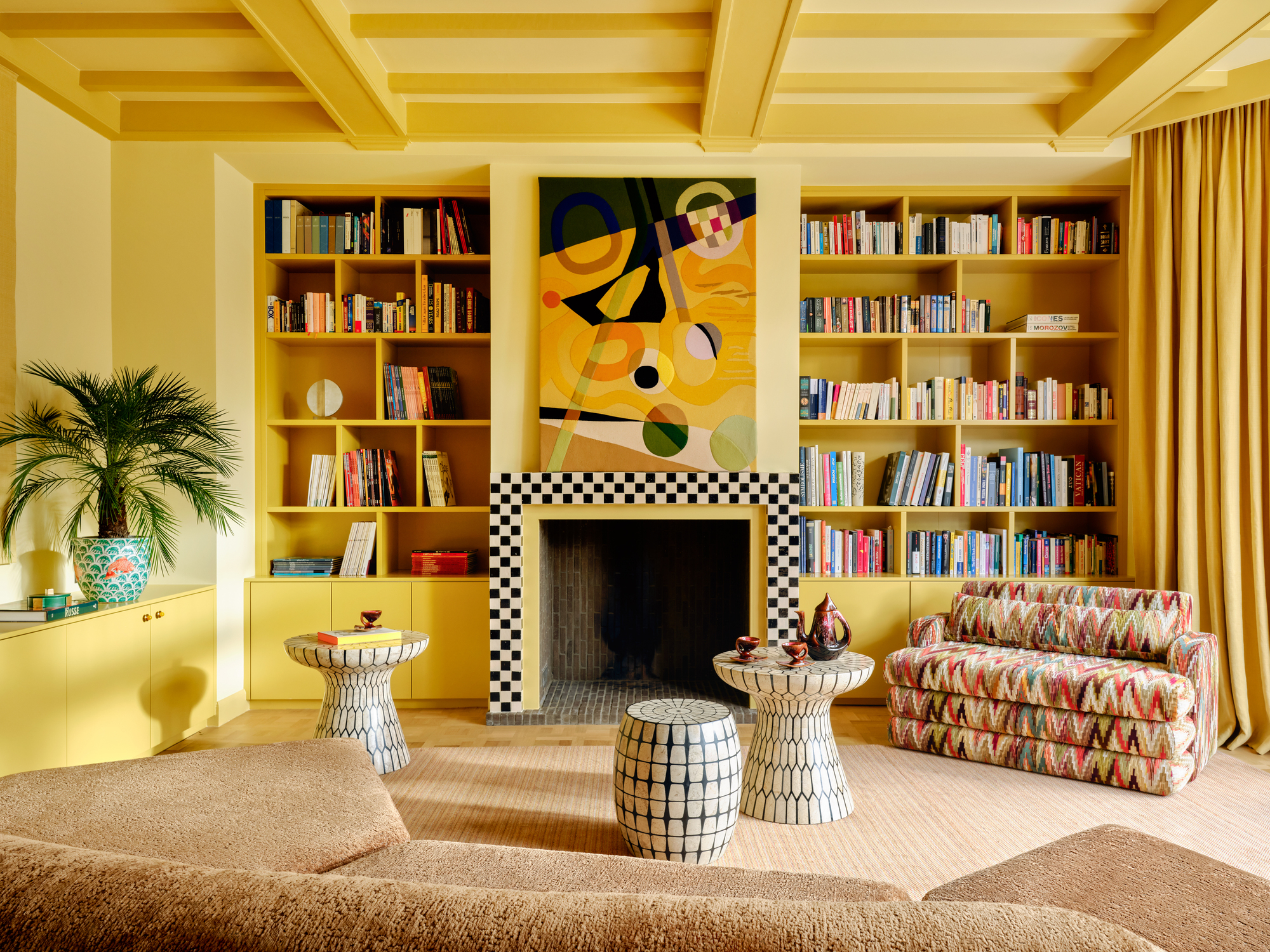 color-drenched yellow living room with painted walls and coffered ceiling, with a built-in bookcase, checkered fireplace, side tables, patterned sofa, console with potted plant