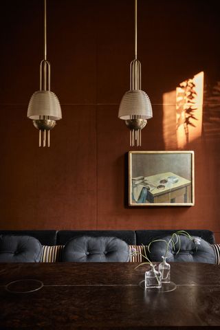 Image of a dining room table that is dark brown and the bench seat is dark blue upholstery with gray-blue cushions. The walls are a black-orange color and there are two gold, metal pendant lights hanging above the table.