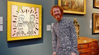 The artist Mr Doodle, a man with ginger hair and wearing a doodle-covered shirt and trousers, stands in front of a canvas of his work showing two figures with lots of expressive lines and a painted frame. He is in an art gallery