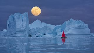 Red sailboat sailing among floating icebergs in front of the full moon rising at Arctic Ocean in Greenland. Sailing through enormously huge icebergs near Ilulissat Icefjord front of the full moon rising. Arctic iceberg reflection in the calm Arctic Ocean waters. Sail boat with red sails cruising among icebergs during midnight sun season. Ship sailing past majestic icebergs during the twilight under a full moon at the blue Arctic Ocean, Ilulissat Icefjord, Ilulissat, Disko Bay, Greenland, Unesco World Heritage Site