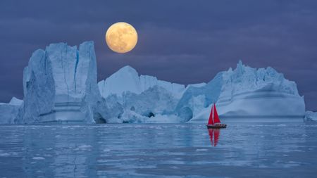 Red sailboat sailing among floating icebergs in front of the full moon rising at Arctic Ocean in Greenland. Sailing through enormously huge icebergs near Ilulissat Icefjord front of the full moon rising. Arctic iceberg reflection in the calm Arctic Ocean waters. Sail boat with red sails cruising among icebergs during midnight sun season. Ship sailing past majestic icebergs during the twilight under a full moon at the blue Arctic Ocean, Ilulissat Icefjord, Ilulissat, Disko Bay, Greenland, Unesco World Heritage Site