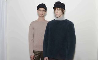 Two male models wearing looks from Gucci's collection. One model is wearing a beige sweater and dark sage coloured pants and the other is wearing a blue jumper with light grey turtle neck jumper underneath. Both models are wearing hats