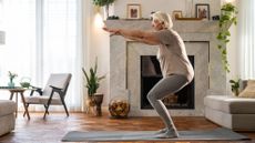 A woman practices squats in a living room on a yoga mat. Her knees are gently bent, arms stretched in front, behind her we see an elegant fireplace and trailing plant.