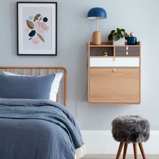 Blue bedroom with double bed, blue duvet, wooden wall mounted side table next to fluffy stool