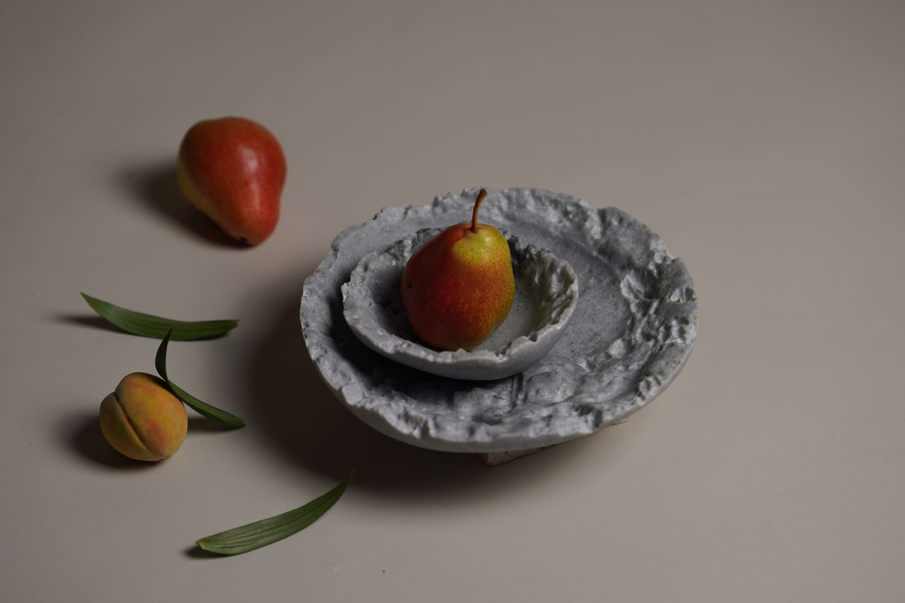 Two organic-shaped, rough grey bowls contain a red pear sitting on a grey surface next to another pear and an apricot.