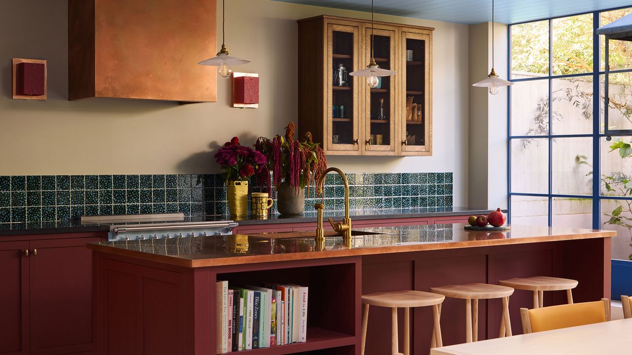 A detailed view of a Tom Morris kitchen showcasing a large red island with a copper countertop and light wood stools. Gold fixtures, a teal tiled backsplash, and an arrangement of deep purple and red flowers create a rich, layered aesthetic against pale yellow walls.