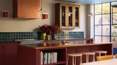 A detailed view of a Tom Morris kitchen showcasing a large red island with a copper countertop and light wood stools. Gold fixtures, a teal tiled backsplash, and an arrangement of deep purple and red flowers create a rich, layered aesthetic against pale yellow walls.