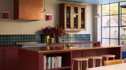 A detailed view of a Tom Morris kitchen showcasing a large red island with a copper countertop and light wood stools. Gold fixtures, a teal tiled backsplash, and an arrangement of deep purple and red flowers create a rich, layered aesthetic against pale yellow walls.
