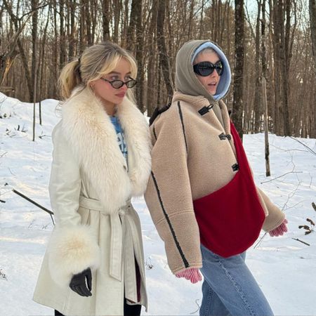 Sabrina Carpenter wears a white fur-trimmed coat over a blue t-shirt, standing next to Emma Chamberlin who wears a borge jacket with a khaki hood.