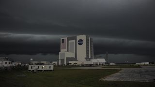 A giant, white, cube shaped buidling with a NASA logo and american flag. Dark grey clouds loom overhead as a storm rolls in.