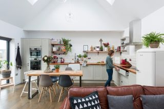 A double-height kitchen diner extension with rooflights, built-in white kitchen units with wooden countertops, and a wooden family dining table, with a woman working at the hob