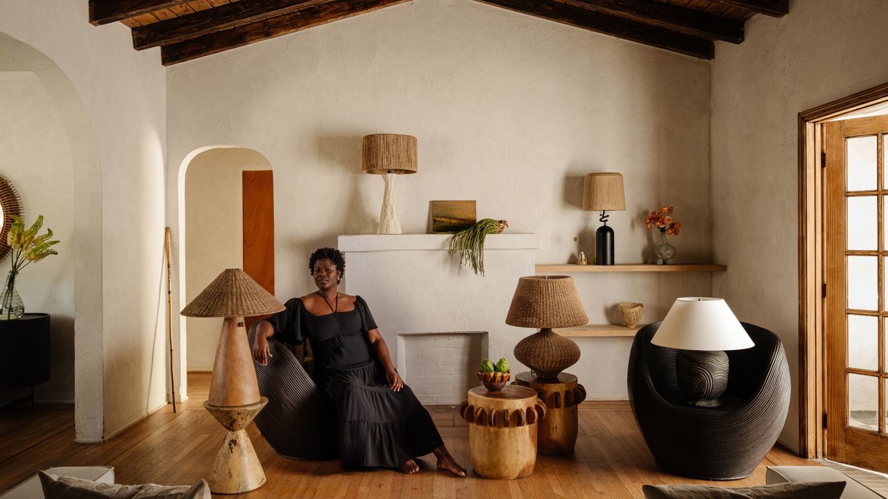 A woman in a black dress poses gracefully sitting on a minimalist armchair in black in a room beautifully decorated with rattan, wood, and glass lamps and homeware.