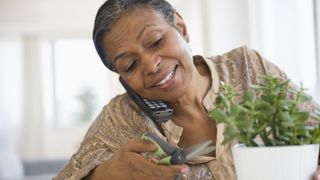 Woman holding a cordless phone to her ear and trimming a house plant in front of her.