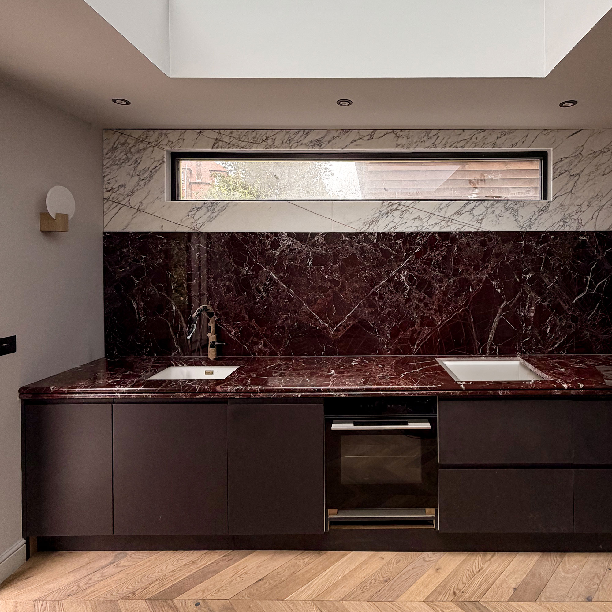 Newly finished kitchen with wooden chevron flooring, red marble worktops and splashbacks, topped with white marble. Dark red cabinetry. High narrow window.