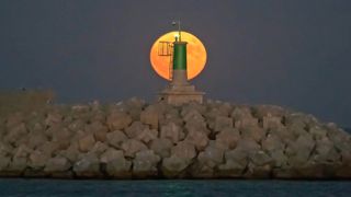 august 2023 full moon in the background behind a green and white lighthouse with water below.