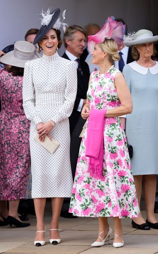 Princess Kate wearing a polka dot dress and black hat with a feather laughing and standing next to Duchess Sophie in a white dress with pink flowers holding a pink scarf over her arm