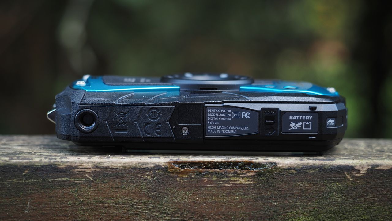Close up of a Pentax WG-90 sitting on a wooden surface in front of woodland