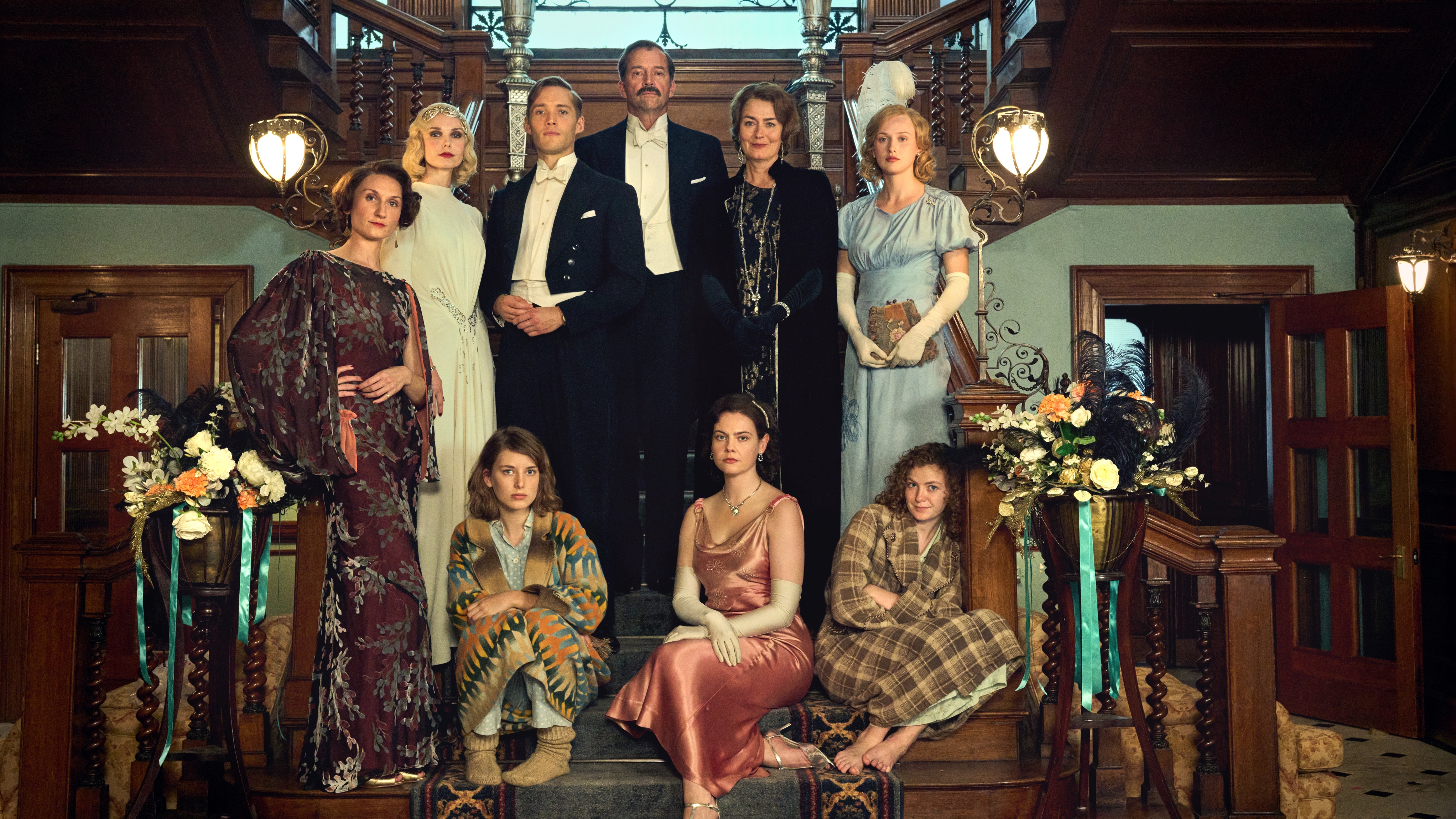 The Mitford family stand together on the staircase of a country house for a family picture, in their eveningwear. In the back row from left to right are: Diana Mitford (Joanna Vanderham), Tom Mitford (Toby Regbo), Farve (James Purefoy), Muv (Anna Chancellor) and Unity Mitford (Shannon Watson). Nancy Mitford (Bessie Carter) stands alone in the middle row, leaning against a bannister and standing just in front of Diana. Seated on the lower row from left to right are Jessica Mitford (Zoe Brough), Pamela Mitford (Isobel Jesper Jones) and Deborah Mitford (Orla Hill).