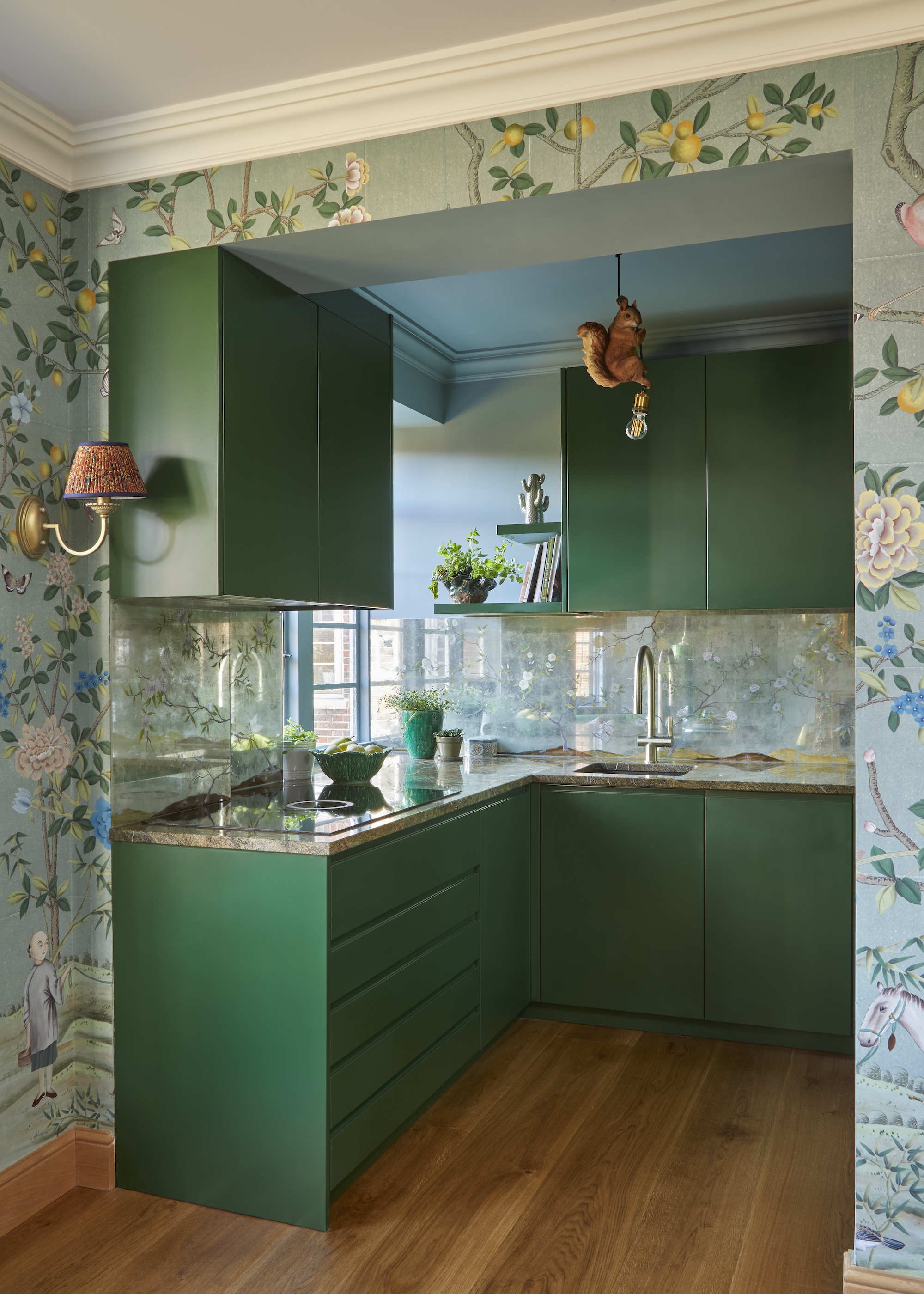 A green kitchen with upper and lower cabinets, the kitchen also has light green wallpaper around it with flowers and fruits painted alongside it.