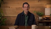 A man with glasses, wearing a dark jacket over a green shirt, sits at a desk in front of a wooden wall background with house plants, a mug, notebooks, and a lamp. 