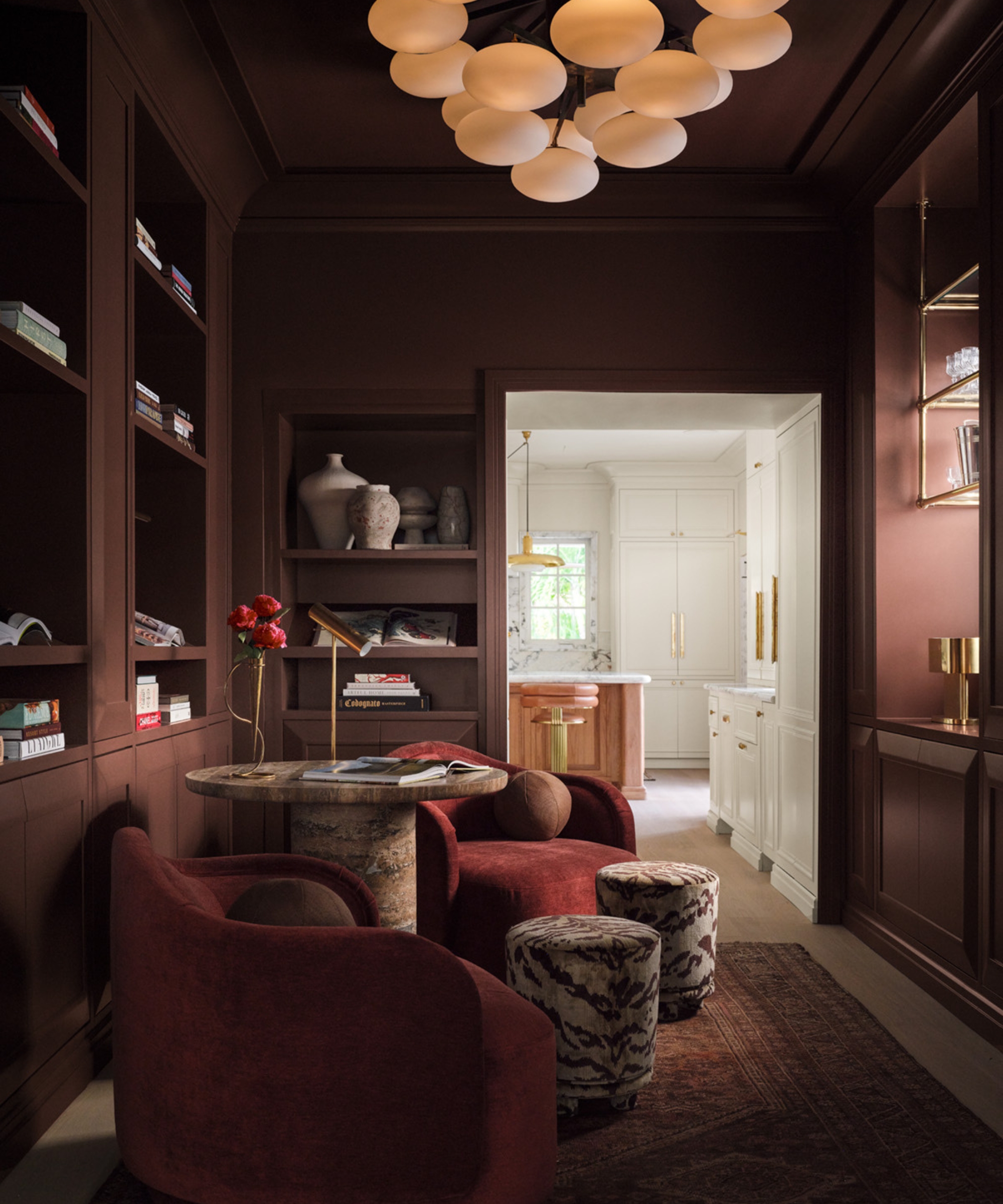 Oxblood-painted library with color-drenched walls, ceiling and shelving, burgundy chairs, patterned stools, and brass lighting.