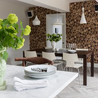 A light grey dining room with log pile print wallpaper, a dark wooden table and white chairs