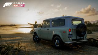 A truck on the beach at sunrise, with a person standing in front with their arms opened wide.