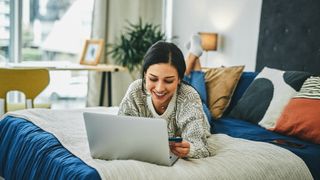 A woman with long dark hair smiles as she buys a mattress online via her laptop during memorial day sales while lying on her current bed which needs replacing