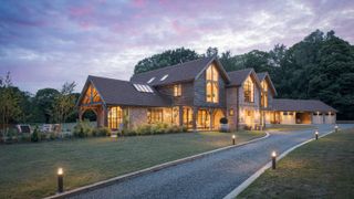 A large oak frame self build shown at dusk. In the foreground there is a long driveway, illuminated by lighting, and a lawned garden. In the background, a woodland.