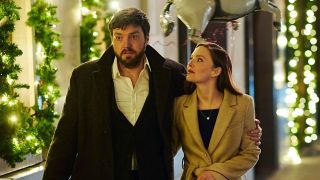 Tom Burke as Cormoran Strike and Holliday Grainger as Robin Ellacott walk arm in arm along a festive high street