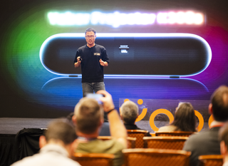 A man in a black t-shirt speaking to an audience at a convention centre. Behind him is a picture of one of the new projectors. One of the people in the audience is out of focus but can be seen taking a picture of the speaker.