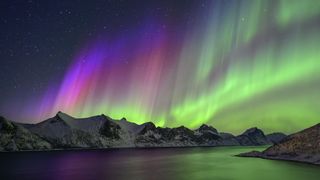 northern lights above mountains and water below. The sky is colored by colorful ribbons of light dancing along, they are mainly green with some purple and red on the left.