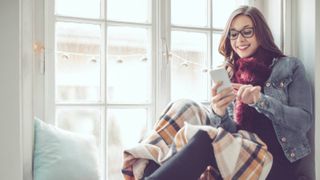 woman sat on window seat in front of multi paned casement windows that are misted over, looking at phone, 