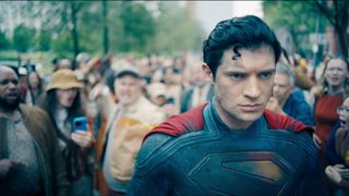 Still from the movie Superman (2025). Superman (man wearing a blue bodysuit with a giant red S on his chest and a red cape) walking through a crowd of angry people.