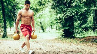 Man walking through a park holding two kettlebells performing a farmer&#039;s walk