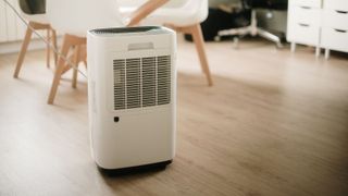 A white dehumidifier with a vented front sits on a light wood floor in a modern room with a dining table, chairs, and storage units in the background.