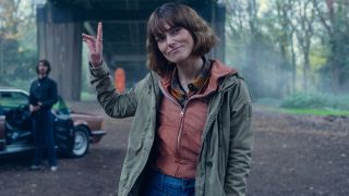 Keira Knightley casually gestures with her hand while Ben Whishaw watches in the background in Black Doves.